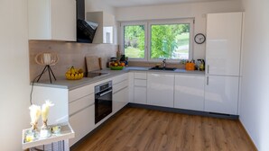 A modern kitchen with white cabinets, a built-in oven, a window with a view of greenery, and a small dining area with a chair and table. - Aura Upper Floor Holiday Home with Lake View, Alpine Panorama, Balconies, Garden & Wi-Fi (Bodman-Ludwigshafen)