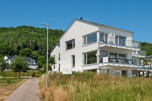 Ein modernes, zweistöckiges Gebäude mit Balkonen, blauem Himmel und einer bewaldeten Hügelkette im Hintergrund.