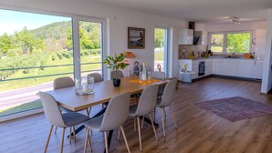Une cuisine moderne avec des meubles blancs, un coin repas doté d’une table et de chaises en bois, et de grandes fenêtres offrant une vue sur un paysage verdoyant.