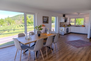 A modern kitchen with white cabinets, a dining area with a wooden table and chairs, and large windows offering a view of a green landscape.