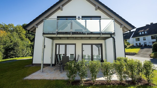 Uma casa moderna de dois andares, com varanda de vidro, um pátio com mobiliário de exterior e um jardim bem cuidado.