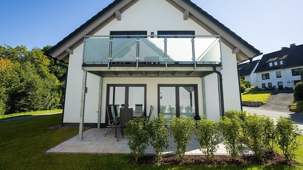 Une maison moderne à deux étages, dotée d’un balcon en verre, d’une terrasse avec mobilier d’extérieur et d’un jardin bien entretenu.