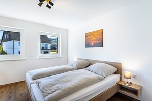 A bedroom with two beds, a nightstand with a lamp, a painting of a sunset, and two windows with views of houses. - Apartment 'Waldhaus' with Mountain View, Balcony and Wi-Fi (Schmallenberg)