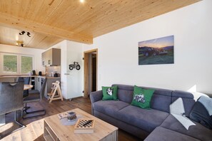 Une cuisine moderne avec un plafond en bois, un coin repas avec une table et des chaises, un canapé gris avec des coussins verts et un tableau représentant un tracteur accroché au mur.
