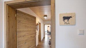 Um corredor com piso de madeira e uma porta à esquerda. Uma placa na parede com o contorno de uma vaca e a palavra 'Muh' é visível.