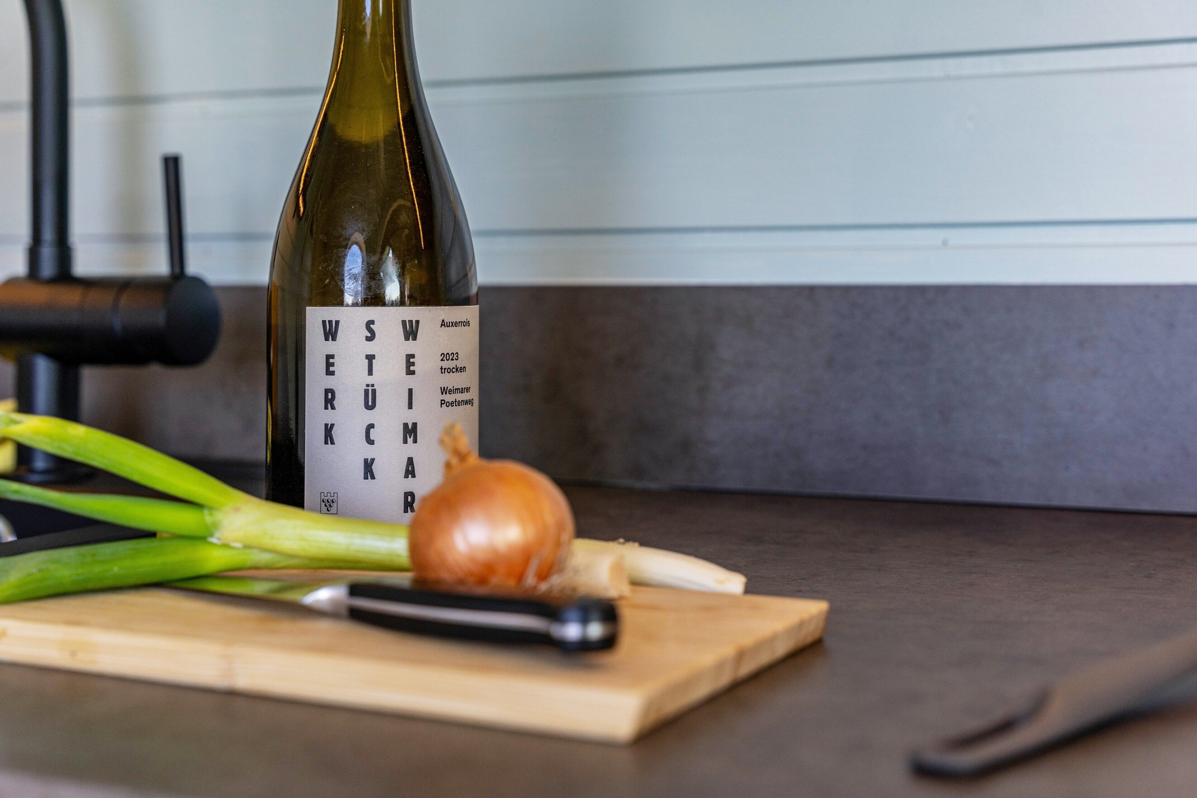 A bottle of wine, a cutting board, a knife, and some vegetables on a kitchen counter.
