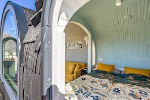 Una habitación con un ventanal grande, una cama con ropa de cama floral, un sillón amarillo y una pared con cuadros.