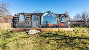 Une petite cabane rustique, dotée d’un toit en pente, d’une grande fenêtre centrale et d’un porche avec deux chaises rouges.