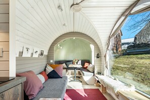 Ein gemütliches Interieur mit gewölbter Decke, einer Sitzecke mit Kissen, einem kleinen Tisch mit einer Vase und einem Fenster mit Blick auf eine Rasenfläche.