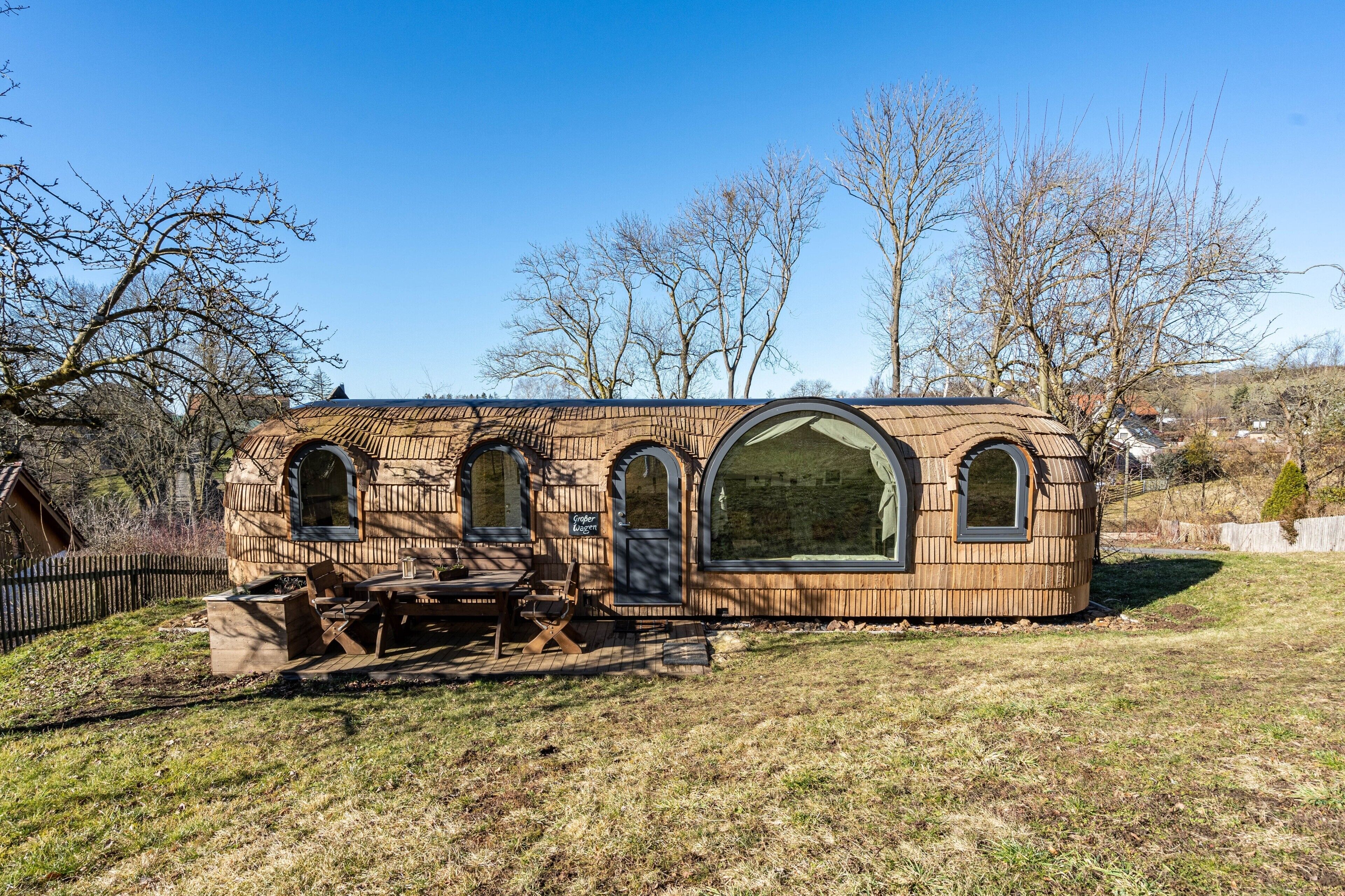 A wooden cabin with arched windows and a large glass door, surrounded by trees and a grassy area.