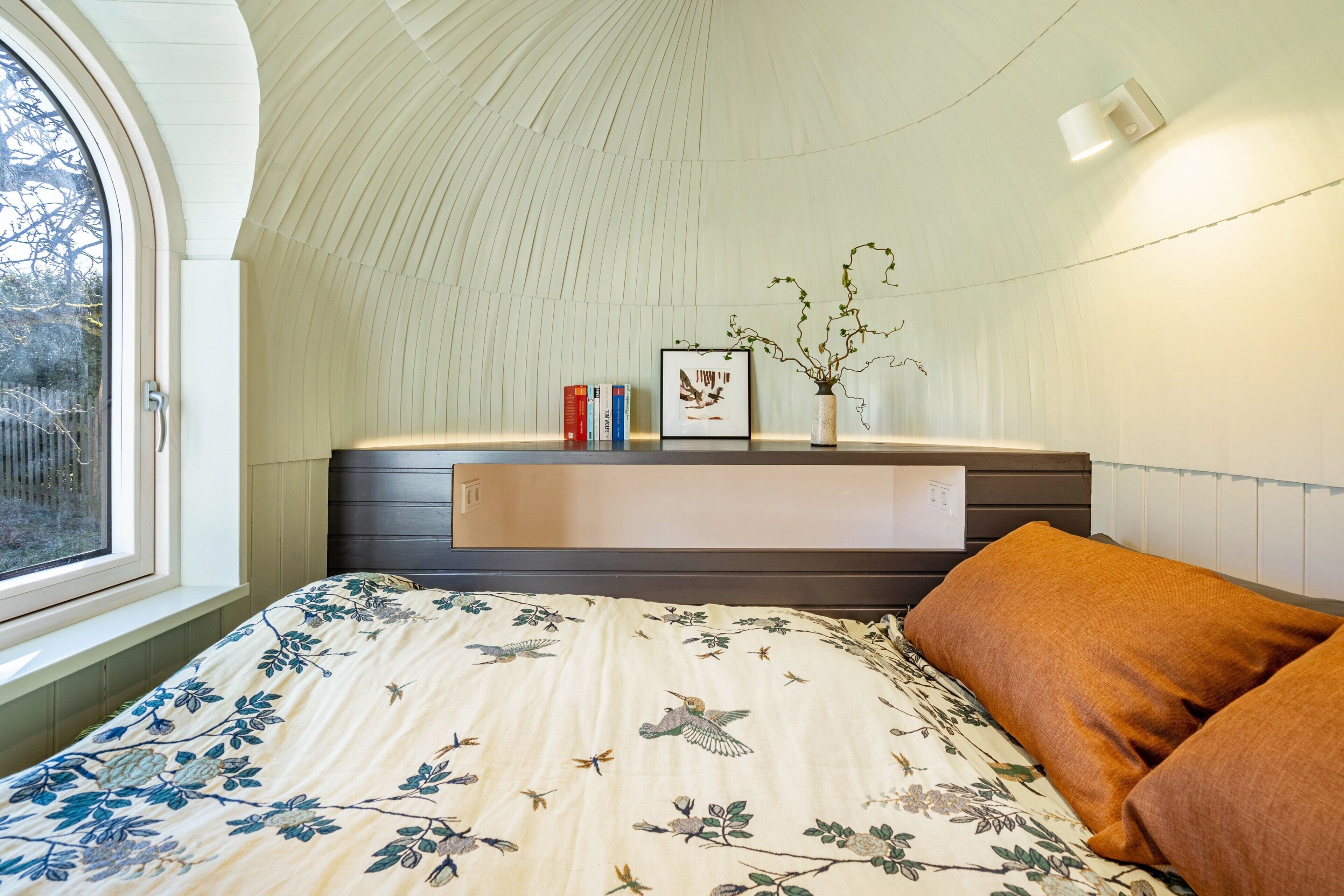 Una camera da letto con soffitto curvo, un letto con biancheria da letto floreale, un comodino con libri e un vaso, e una finestra vista alberi.