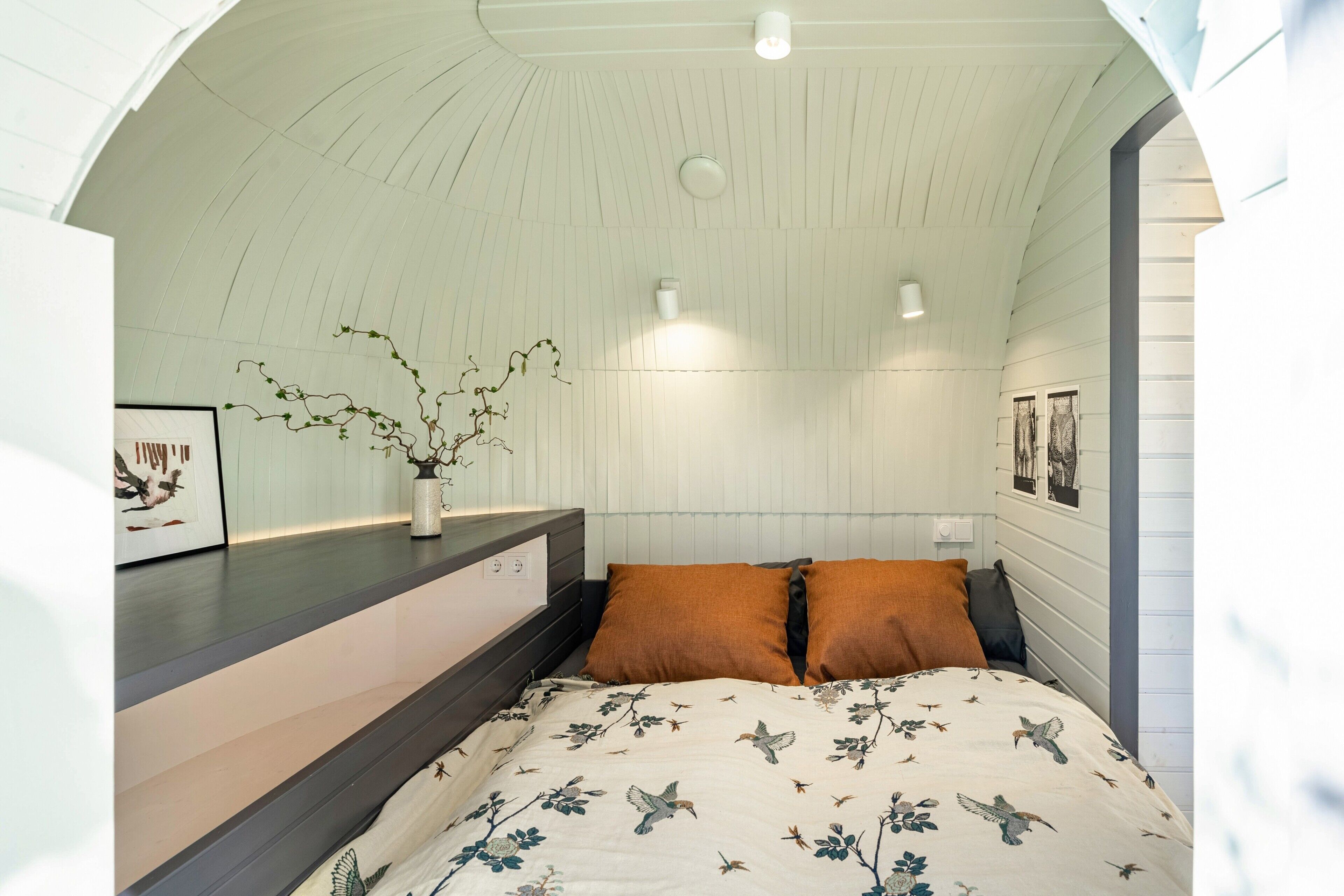 Un dormitorio con techo curvo, una cama con colcha estampada, una mesita de noche con un florero y luces fijadas a la pared.