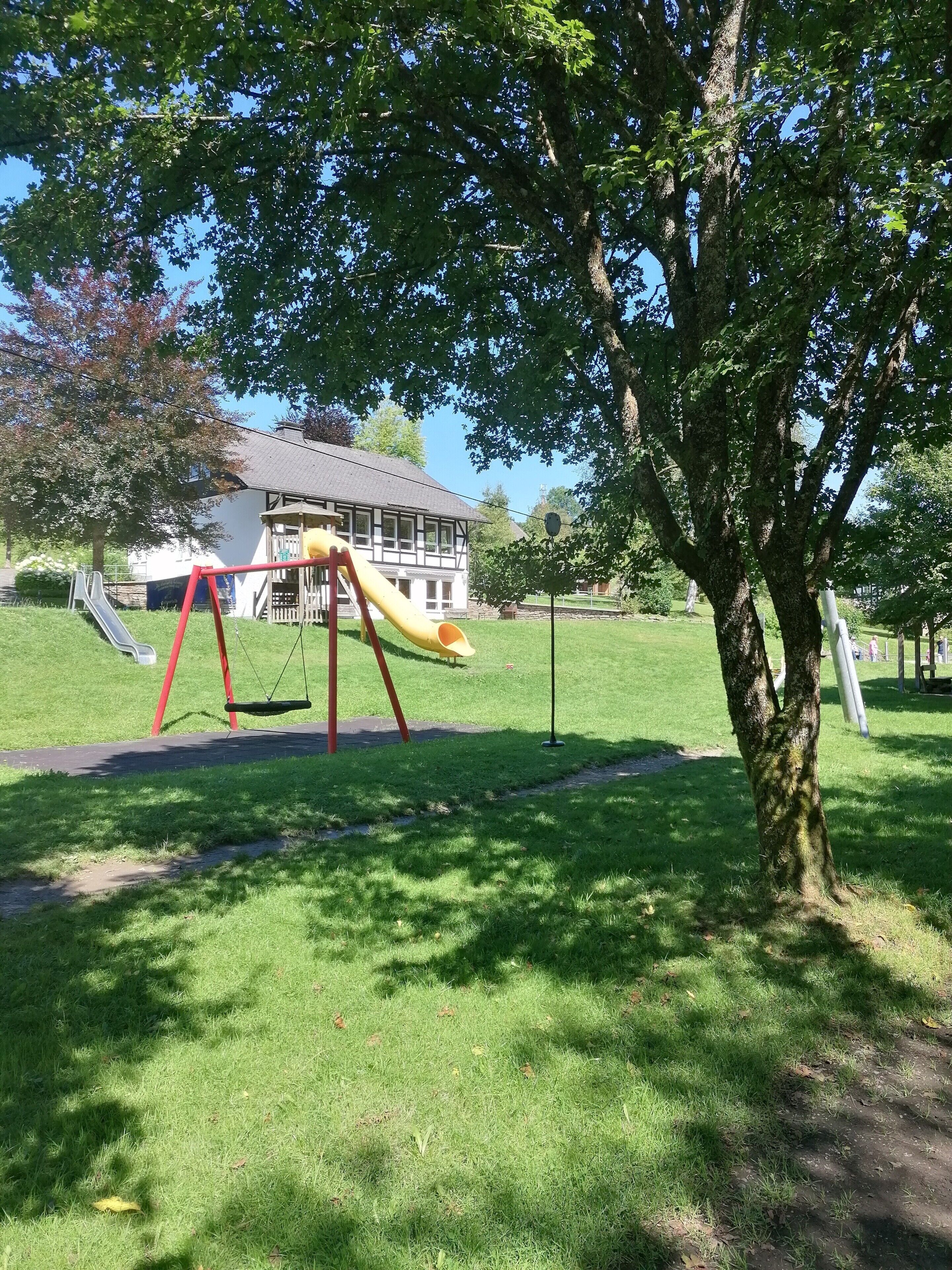 A playground with a yellow slide and swings, a tree, and a building in the background.