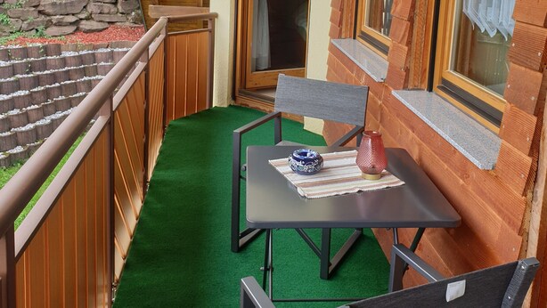 Un balcón de madera con una mesa y sillas, un muro de piedra y vistas a una casa.