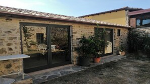 Un bâtiment en pierre doté de grandes portes coulissantes en verre, d’un petit patio avec une table et des chaises, et de plantes en pot.
