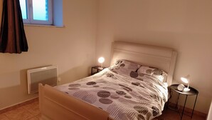 A bedroom with a bed, two bedside tables with lamps, a radiator, a window with curtains, and a patterned bedspread. - Apartment with garden 30min from Lille! (Vieux-Berquin)