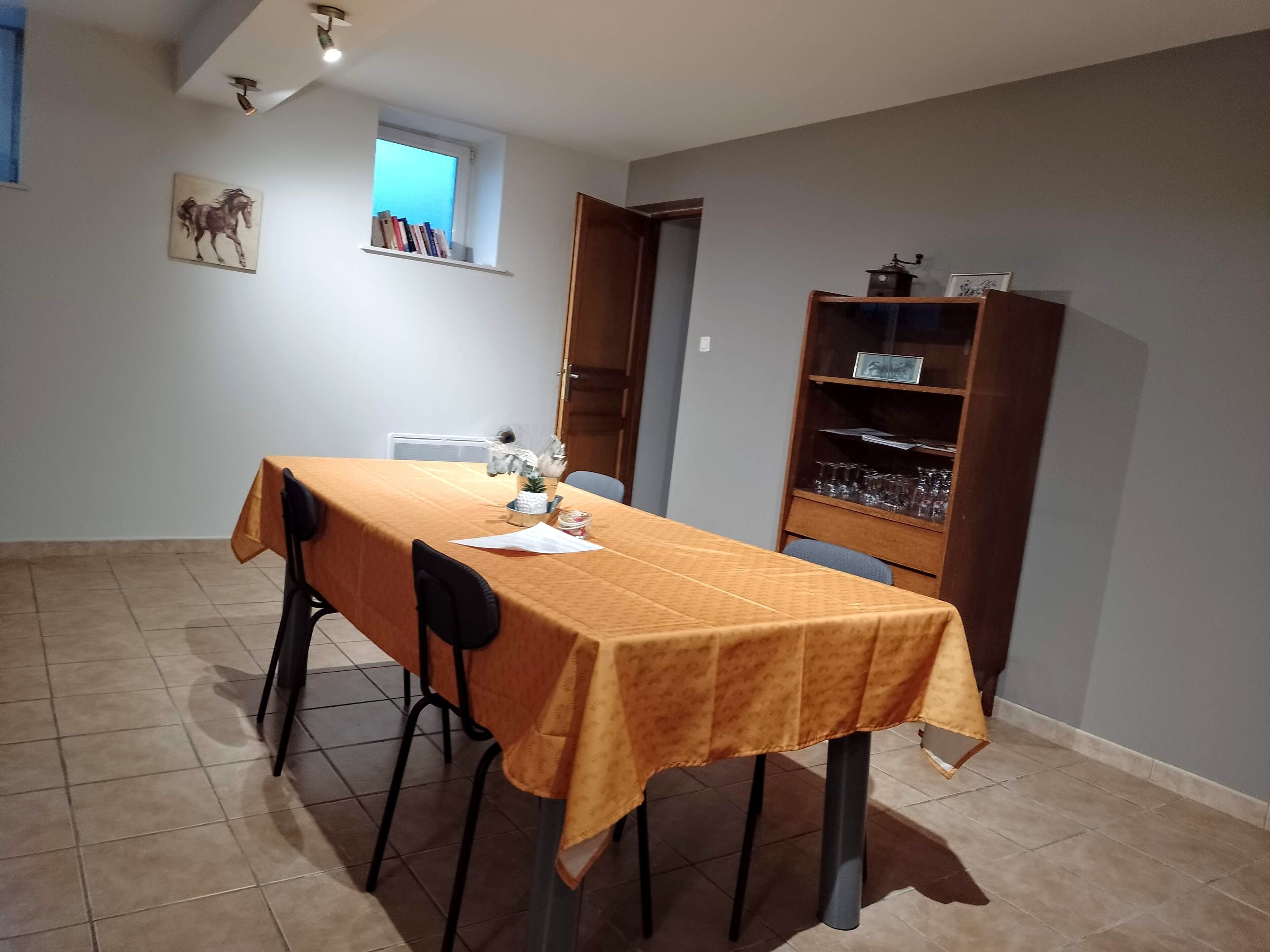 A dining area with a table covered by a yellow tablecloth, chairs, a small shelf with books, and a wooden cabinet with glasses.