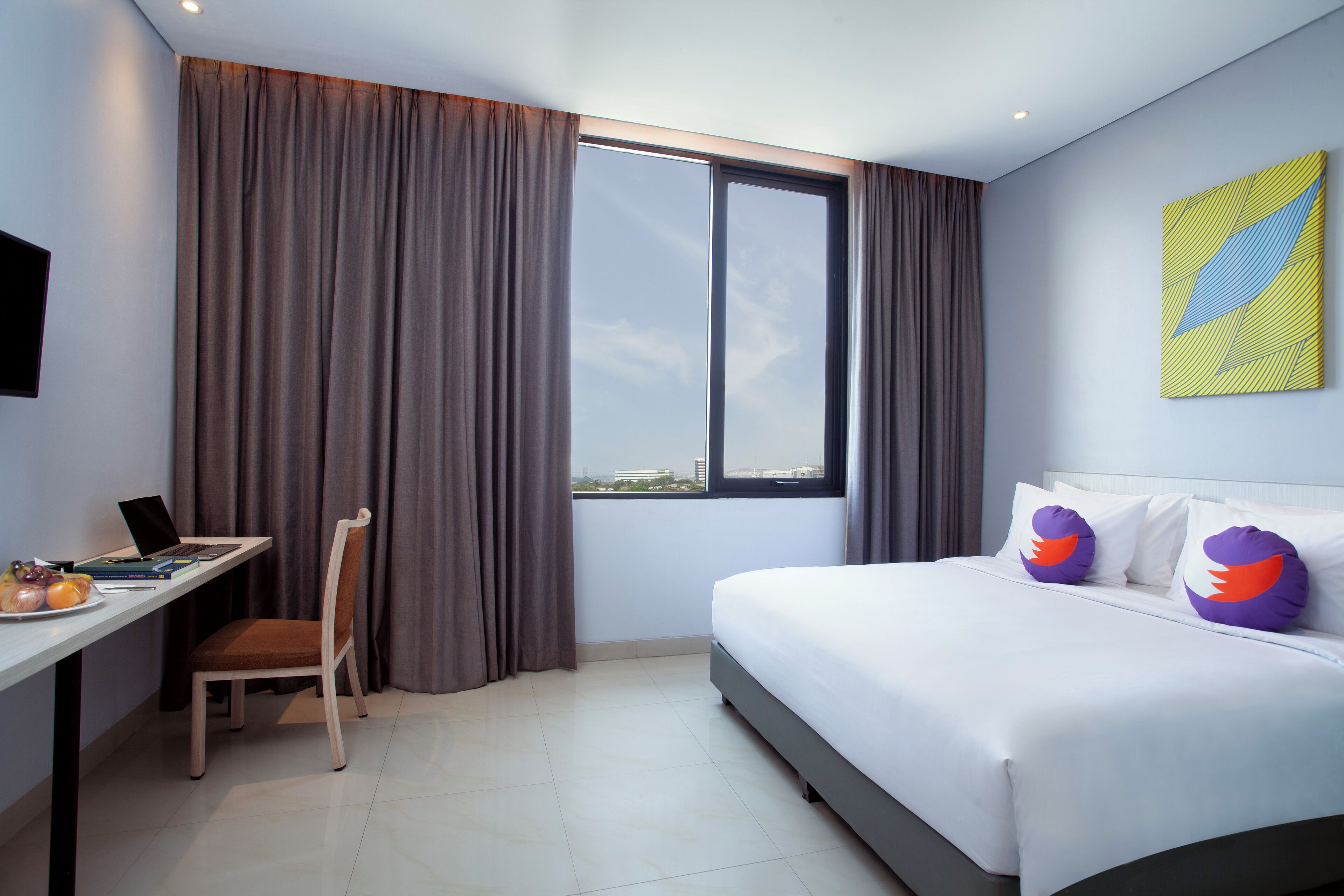 Habitació d'hotel amb llit, escriptori, portàtil, cadira i una finestra gran amb cortines.