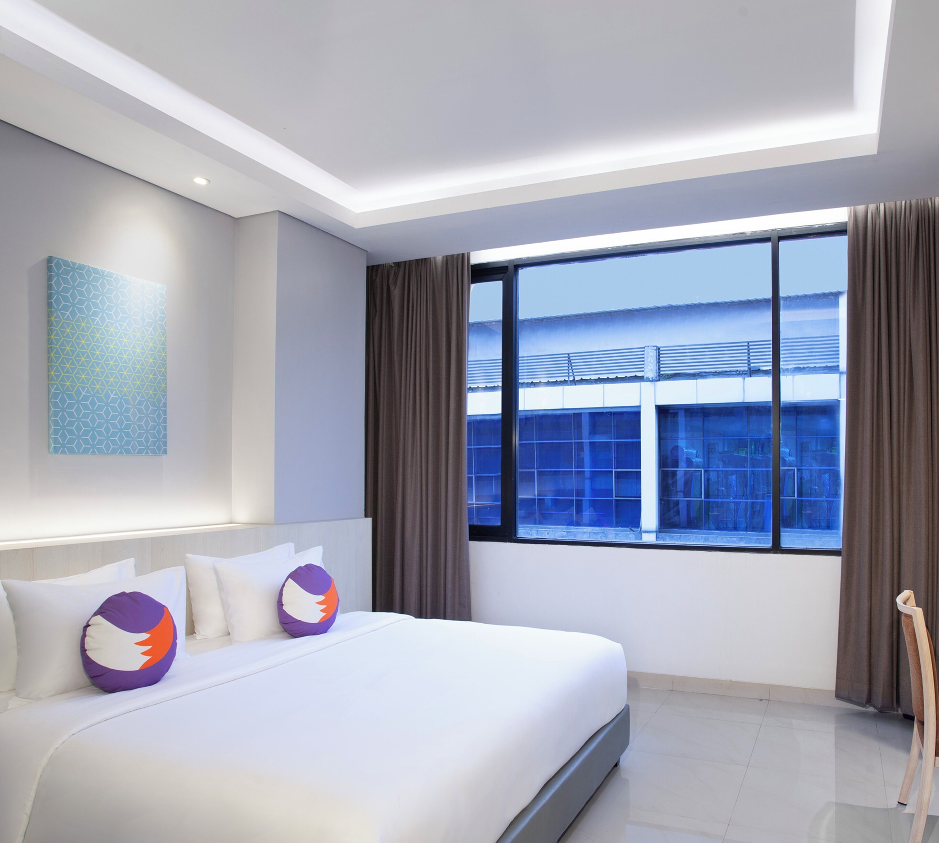A modern hotel room with a large bed, a view of a building through the window, and a painting on the wall.