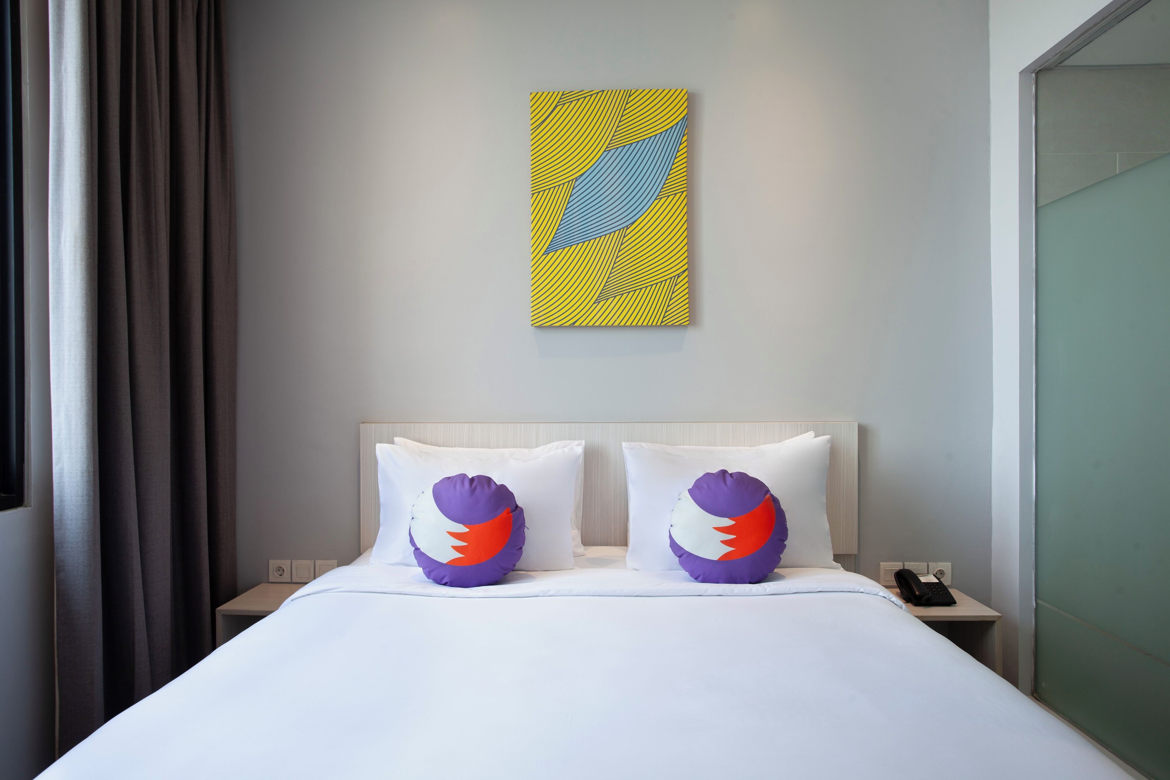 A hotel room with a bed, two decorative pillows, a nightstand, a phone, and a wall art piece.