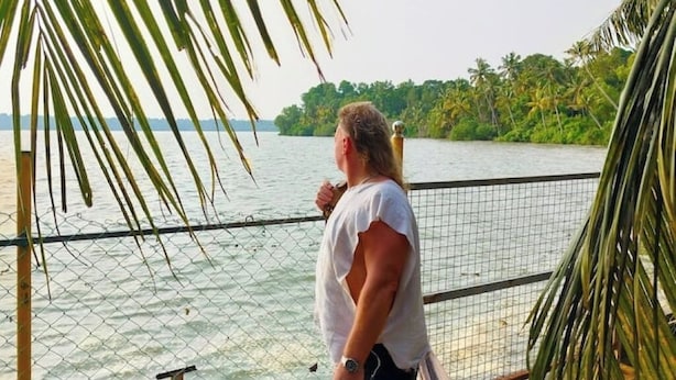 A person standing by a fence overlooking a body of water with palm trees and greenery in the background.