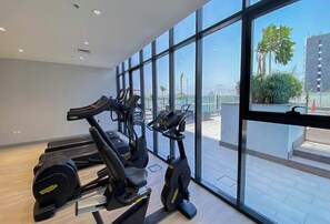 A gym with treadmills and stationary bikes, large windows, and a view of palm trees and buildings. - Bloomfields Studio - Azizi Riviera 19 - Meydan (Dubai)