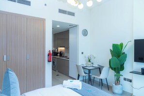 Ein modernes Hotelzimmer mit Bett, Schreibtisch, Stuhl, einer Zimmerpflanze, einem Fernseher und einer durchgehenden Tür erkennbaren Küchenzeile.