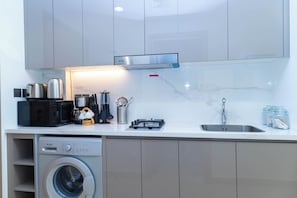 A compact kitchen with a washing machine, microwave, toaster, and a stainless steel range hood. - Bloomfields Studio - Azizi Riviera 19 - Meydan (Dubai)