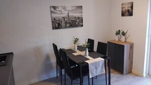 A dining area with a table set for four, two vases with plants, a cityscape painting, and a cabinet with additional plants. - Cozy bungalow for 4 people in the beautiful Hunsrück (Morbach)