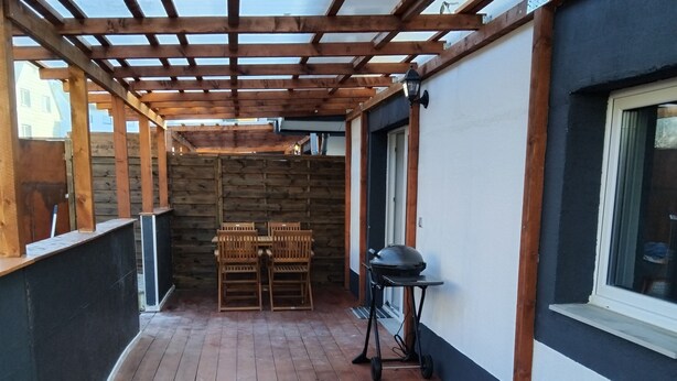 A covered patio with wooden flooring, a seating area with chairs, and a grill. - Cozy bungalow for 4 people in the beautiful Hunsrück (Morbach)