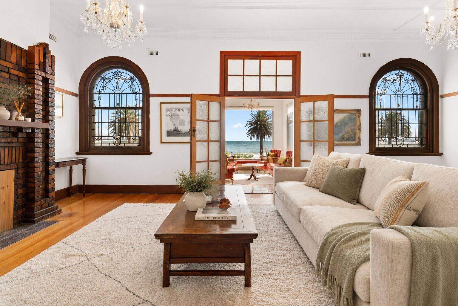 A spacious living room with a large sofa, a coffee table, and a fireplace. There are two large windows with views of the ocean.