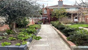 Ein schmaler Betonweg, der von Back- und Steinrändern gesäumt wird und zu einem Wohngebäude mit sichtbarer Treppe und einem verschlossenen Eingang führt.