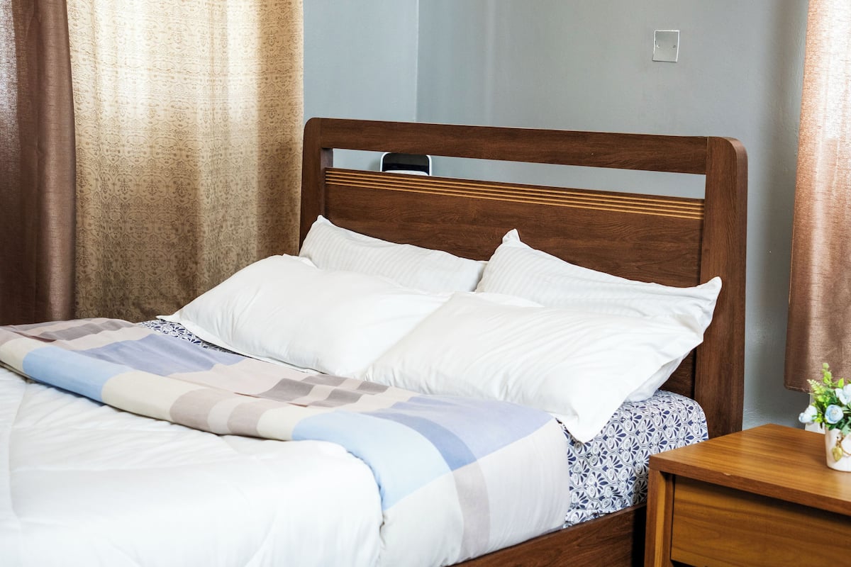 A neatly made bed with white linens and a patterned blanket, a wooden headboard, and a bedside table with a vase of flowers.