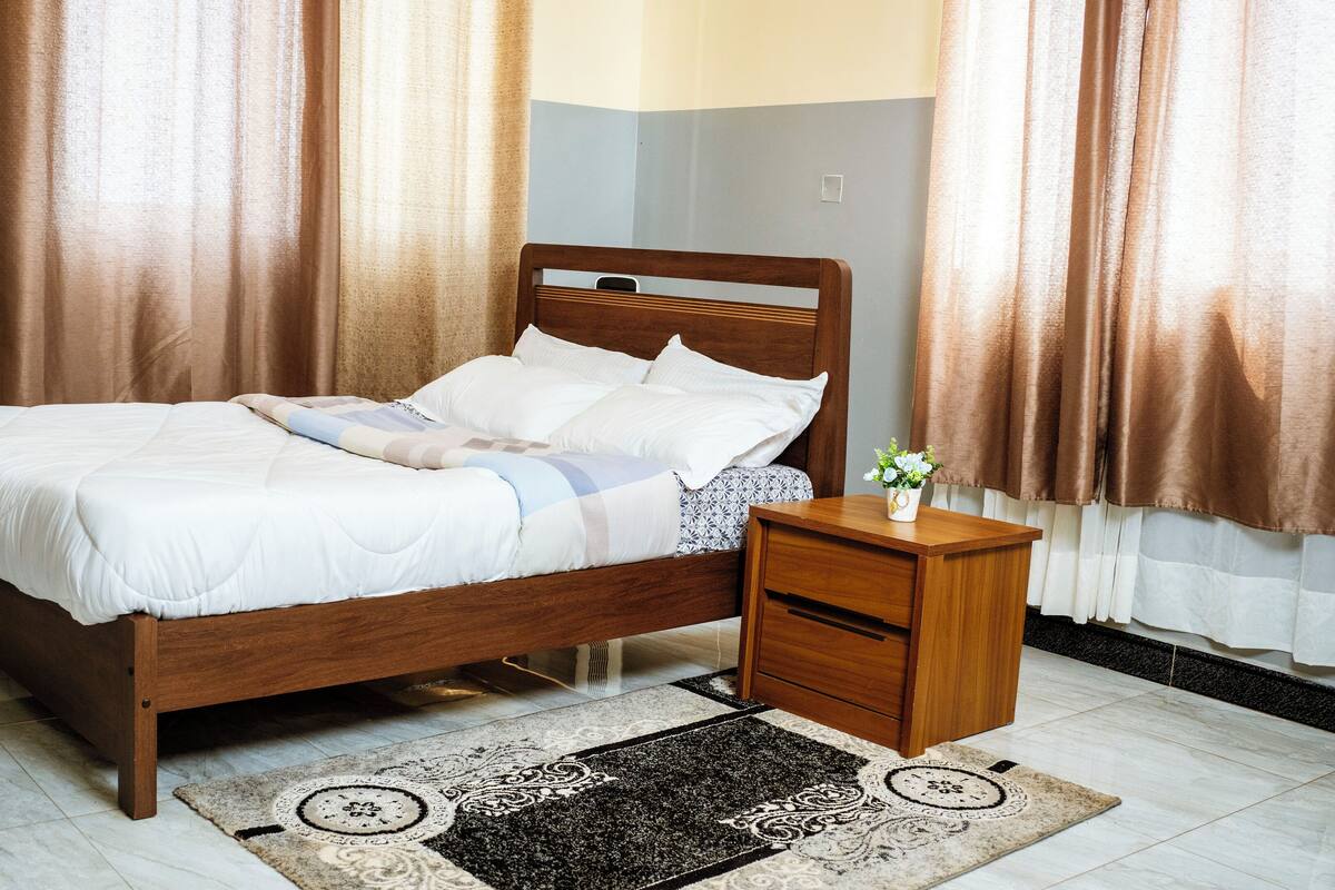 A neatly made bed with white bedding and a blue and white striped pillow, a wooden nightstand with a small potted plant, and a patterned rug on a tiled floor.