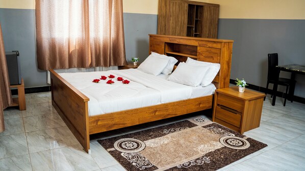 A neatly made bed with white linens and red roses, a wooden nightstand with a vase, a small table with a plant, and a chair. - Northern Gateway Hotel And Lounge (Kamdini)