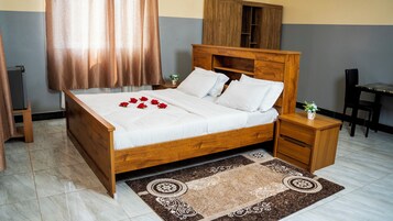Una cama bien hecha con sábanas blancas y rosas rojas, una mesita de noche con un jarrón, una pequeña mesa con una planta y una silla.