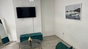 A modern living room with a flat-screen TV mounted on the wall, a round glass coffee table with a metallic vase, teal upholstered seating, and a framed picture of a seascape. - Exquisite 1 Bedroom (Orpington)