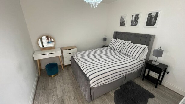 A bedroom with a bed, a desk, a chair, a mirror, a bedside table with a lamp, and a small rug. - Exquisite 1 Bedroom (Orpington)