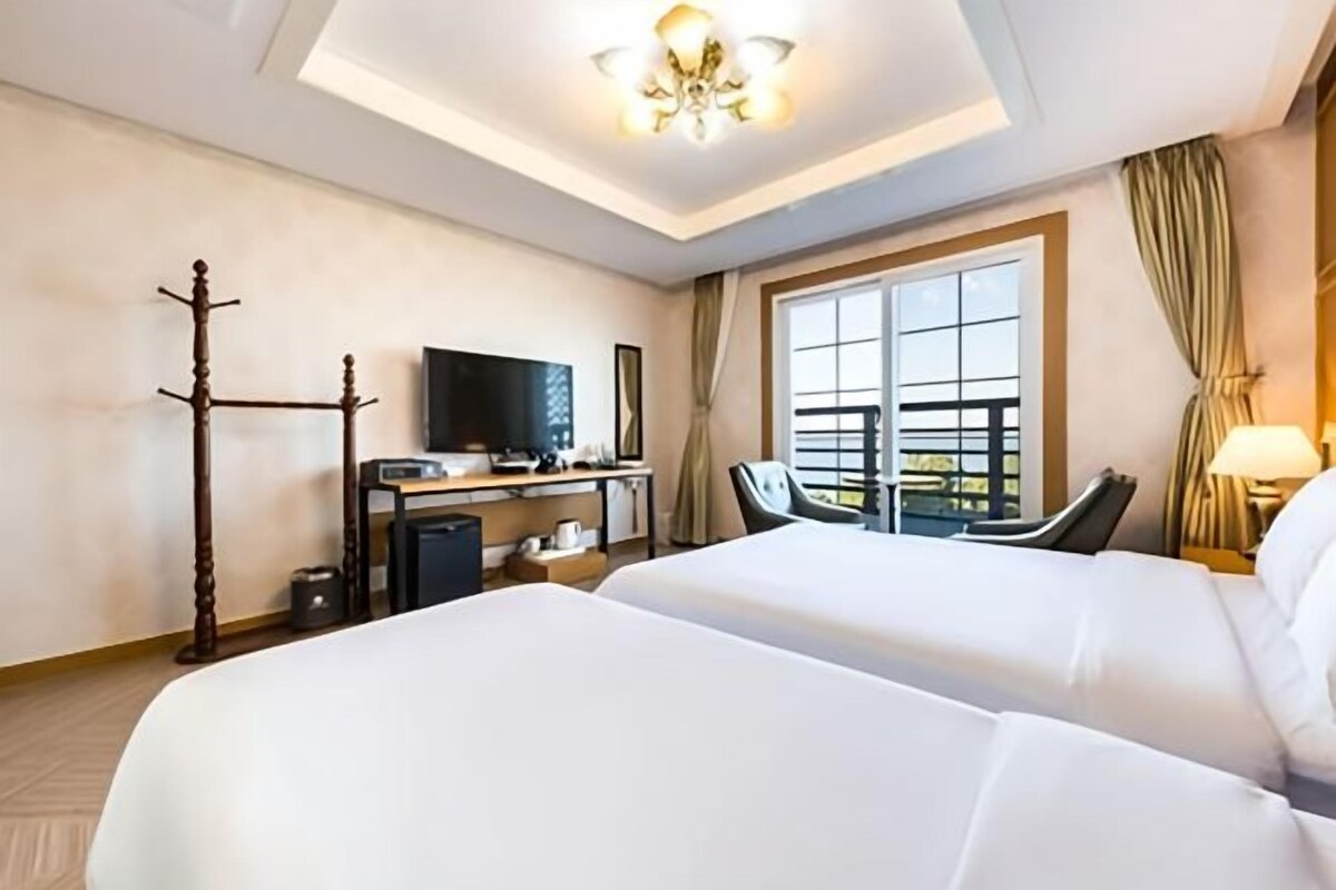 Ein Hotelzimmer mit zwei Betten, einem Schreibtisch mit Fernseher, einem Stuhl, einem Fenster mit Vorhängen und einer Deckenleuchte.