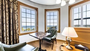 Une chambre d’hôtel à l’agencement incurvé, comprenant une table et deux chaises, une lampe et de grandes fenêtres donnant sur la mer.