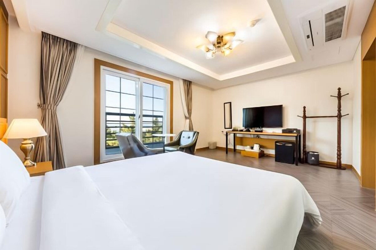 Ein Hotelzimmer mit einem großen Bett, einem Schreibtisch mit Fernseher, einem Sessel und einem Fenster mit Vorhängen.