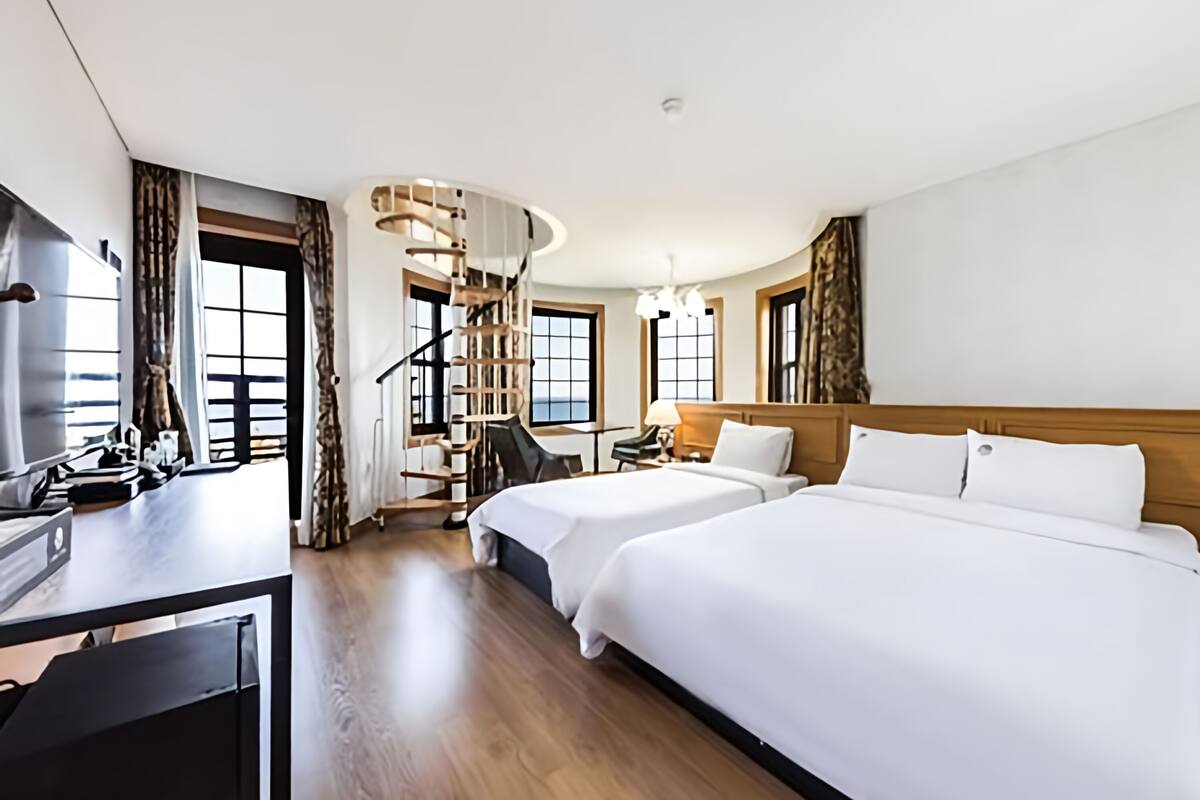 Habitación de hotel con dos camas, un escritorio, una silla, una escalera y ventanas grandes.