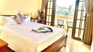 A hotel room with a large bed, a small table with a vase, and a balcony with chairs. - Aman Riverside Hotel (Da Nang)
