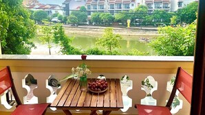 A balcony with a wooden table, two chairs, a vase with flowers, and a bowl of fruit. In the background, there are buildings with red roofs and a body of water. - Aman Riverside Hotel (Da Nang)