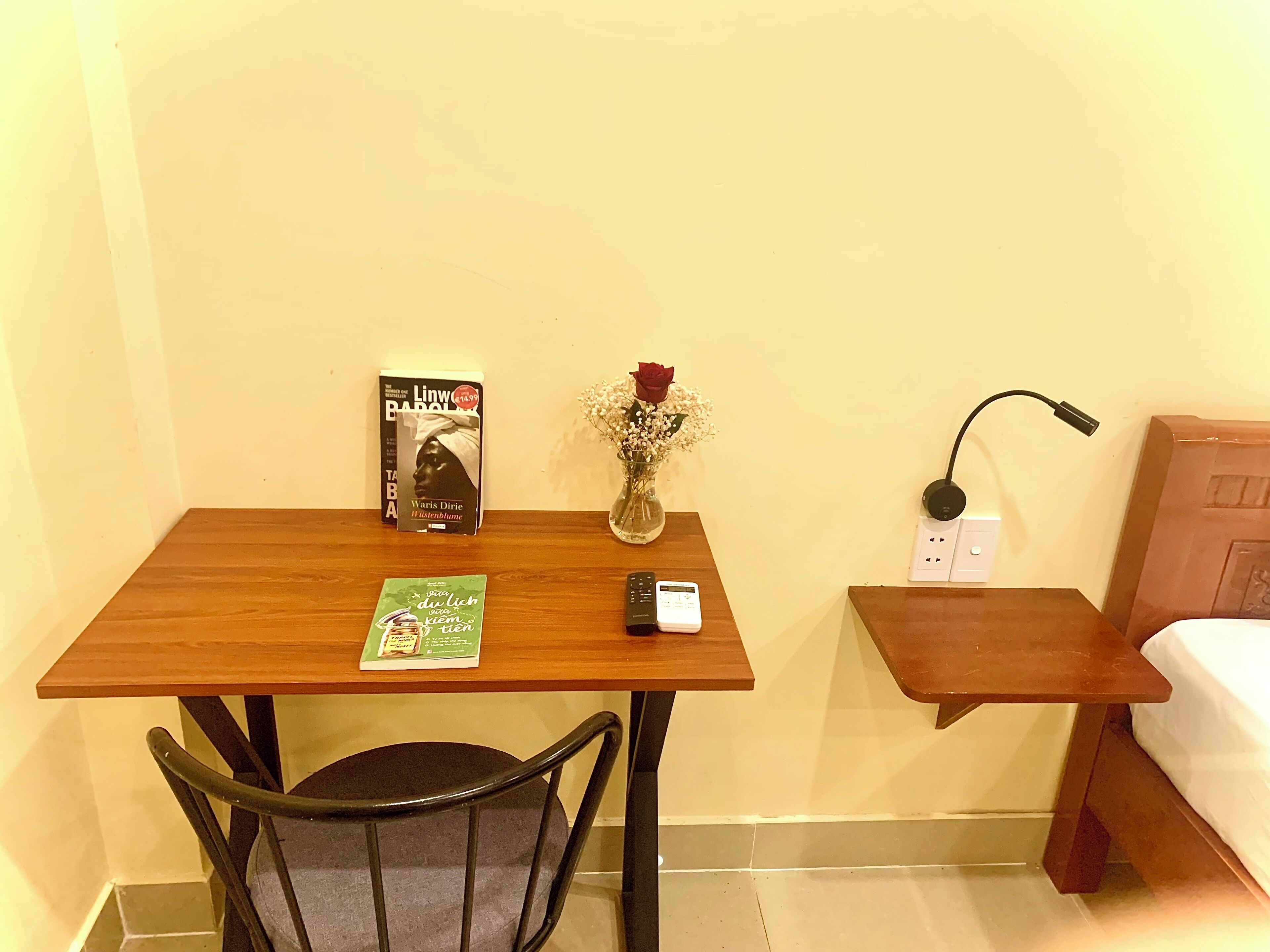 Un pequeño escritorio de madera con una silla, un libro, un florero con flores, una lámpara y un enchufe.