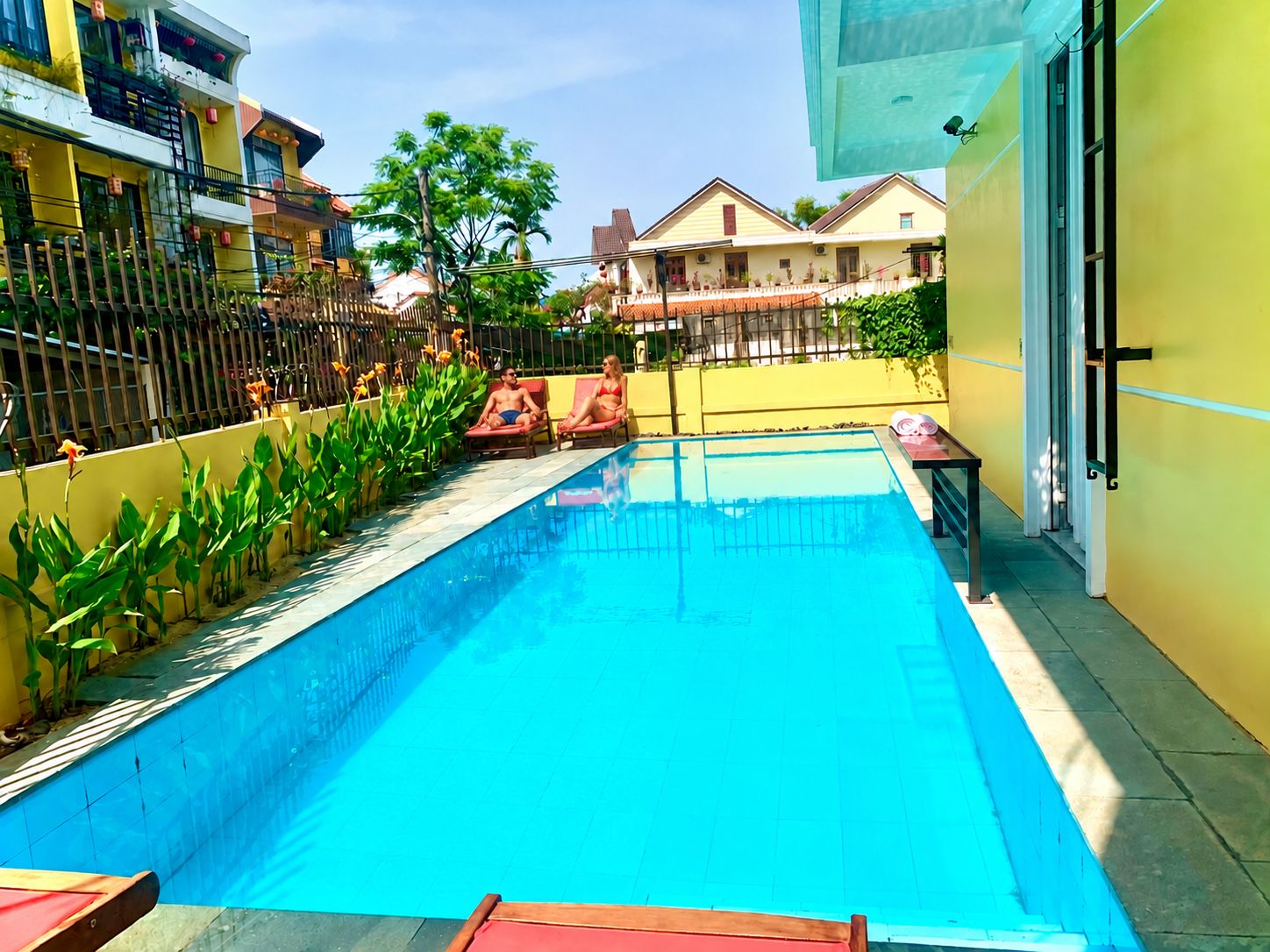 Una piscina con sillones de descanso y una mesa, rodeada por una valla y follaje.