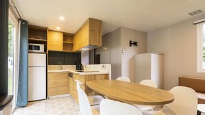 Cocina compacta con armarios de madera, un microondas y un lavamanos. Hay una zona de comedor con una mesa de madera y sillas blancas.