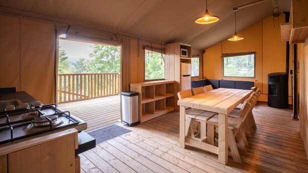A wooden cabin interior with a dining table, benches, a stove, and a fireplace.