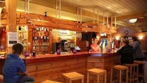 Un espace bar en bois, avec des tabourets, un comptoir et des étagères remplies de bouteilles et de verres.