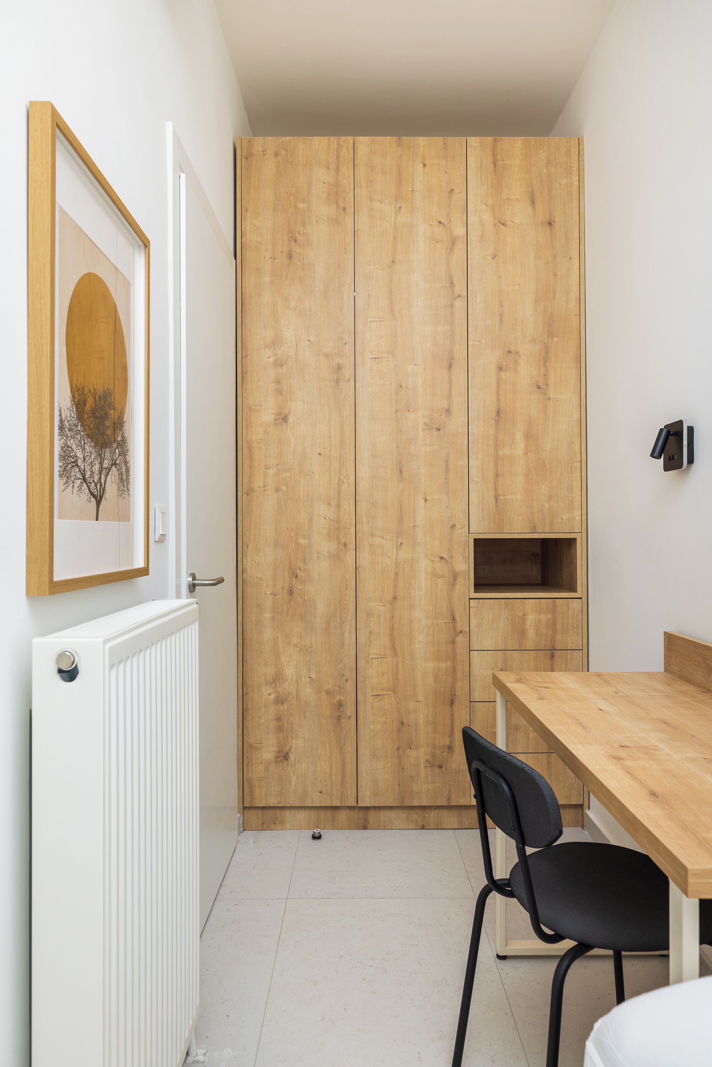 A room with a wooden wardrobe, a white radiator, a framed picture, a wooden table, and a black chair.